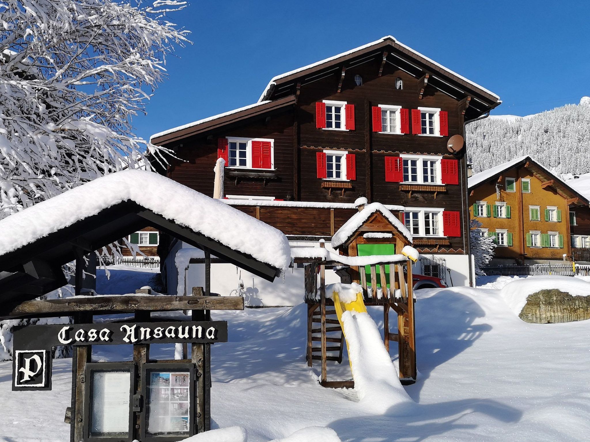 En byggnad av trä med röda fönsterluckor, en skylt med texten ”Casa Ansau”, och en snötäckt lekplats.