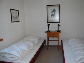 Une chambre simple avec deux lits simples, une petite table en bois avec deux lampes et deux tableaux encadrés accrochés aux murs.
