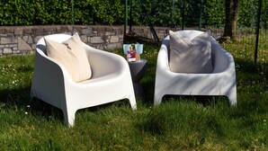 Two white chairs with cushions, a small table with a bottle and a bowl, and a stone wall in the background. - Villa Violetta (Menaggio)