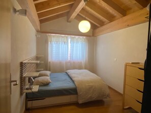 Une petite chambre avec un lit simple, un plafond en bois, une fenêtre avec des rideaux légers, une lampe de chevet et une commode en bois.