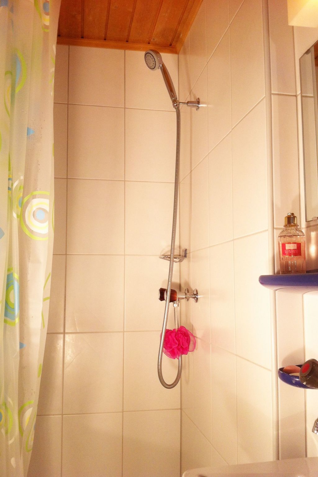 A shower with a handheld showerhead and a wall-mounted faucet, a pink bath sponge, and a bottle of shampoo on a shelf.