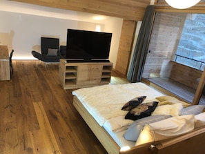 Ein Schlafzimmer mit einem Bett, einem Fernseher auf einem Holztisch und Blick ins Freie durch ein Fenster.