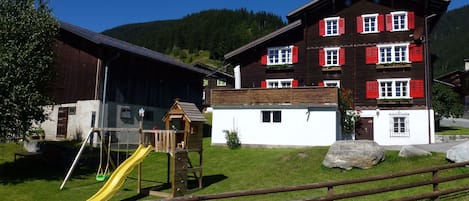 Ein hölzerner Spielplatz mit Rutsche und Schaukeln, ein weißes Gebäude mit roten Fensterläden und ein großer Felsen im Vordergrund.