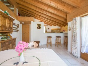 A room with a wooden ceiling, a dining table with a vase of flowers, a kitchen area with stools, and a door leading to another room. - Cuntera Venzin, (Curaglia), 5002 by Interhome (Baselgia)