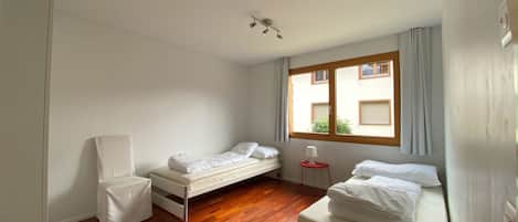 A small, bright room with two single beds, a chair, a small table, and a window with curtains.