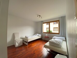 A small, bright room with two single beds, a chair, a small table, and a window with curtains. - Dentervitgs, (Sedrun), by Interhome (Sedrun)