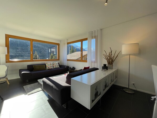 A modern living room with a sofa, a coffee table, and a cabinet. Large windows offer a view of mountains. - Dentervitgs, (Sedrun), by Interhome (Sedrun)