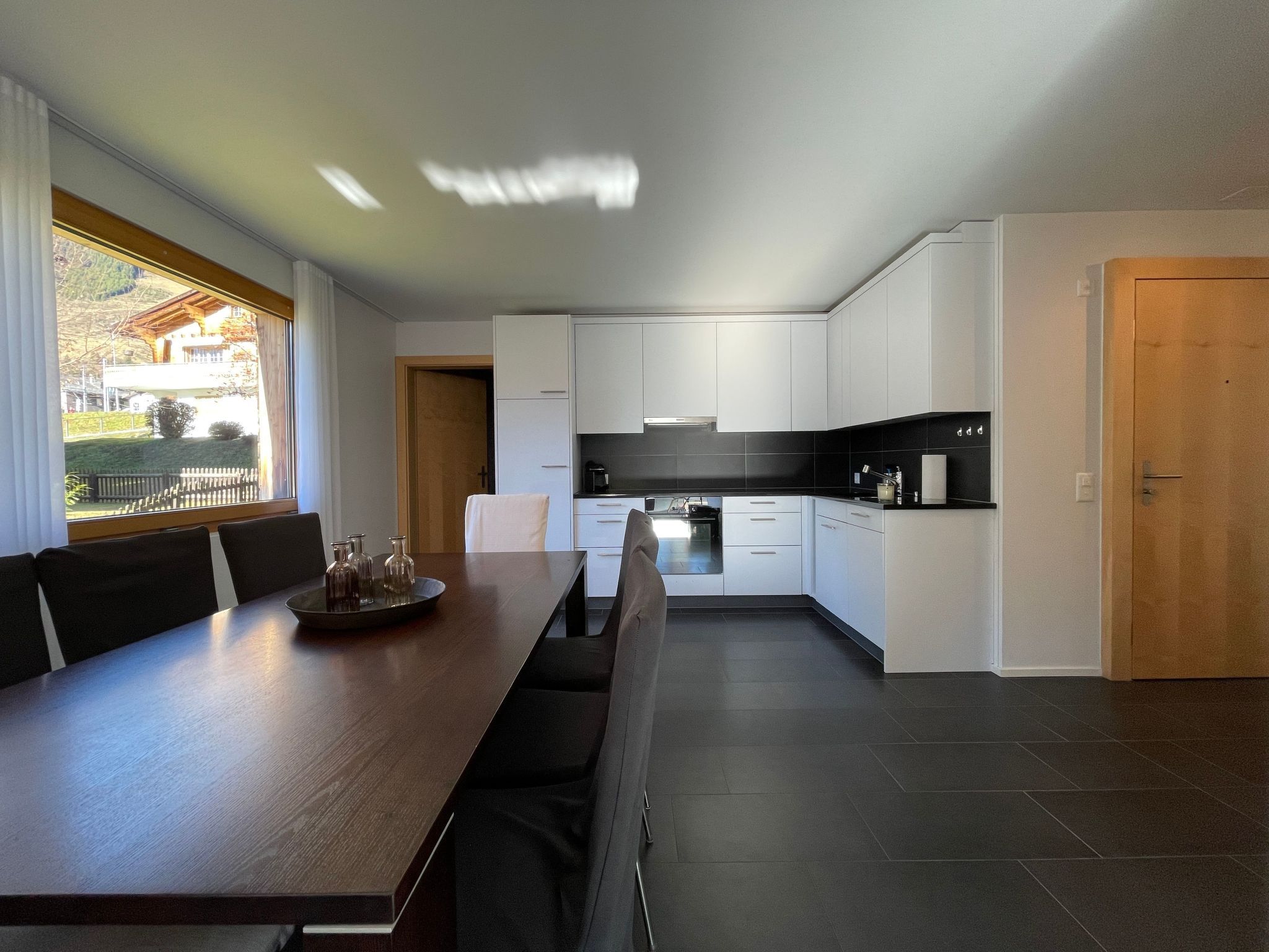 Una cocina moderna con armarios blancos, encimera oscura y un comedor con mesa y sillas de madera.
