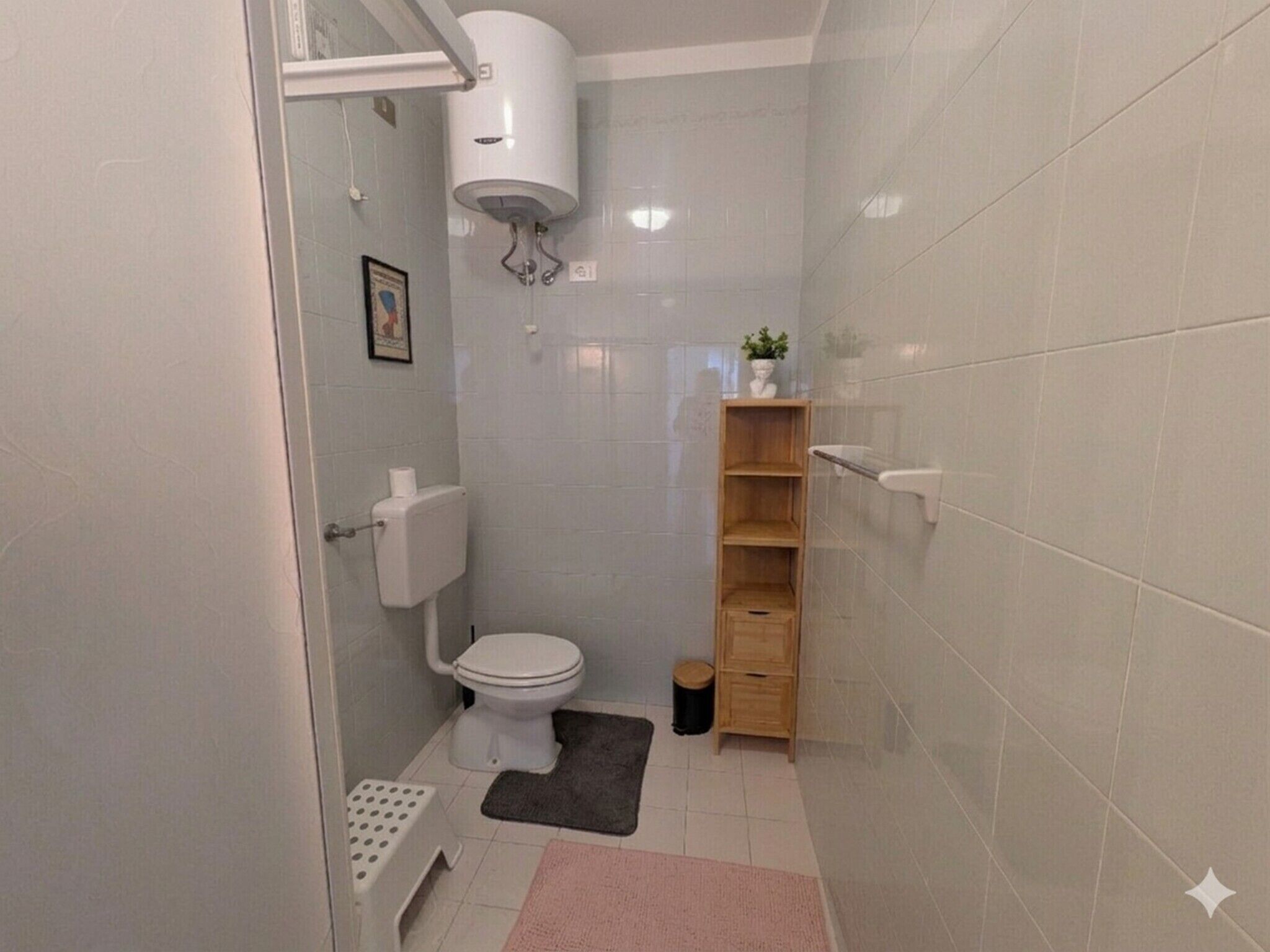 A small, compact bathroom with a toilet, a white sink, a shower area, and a wooden shelf unit.