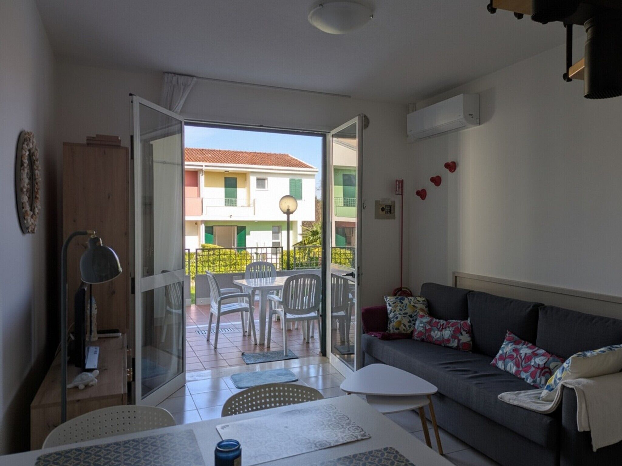 Una sala de estar con un sofá, una mesita, una lámpara y vistas a un balcón con sillas y un edificio al fondo.