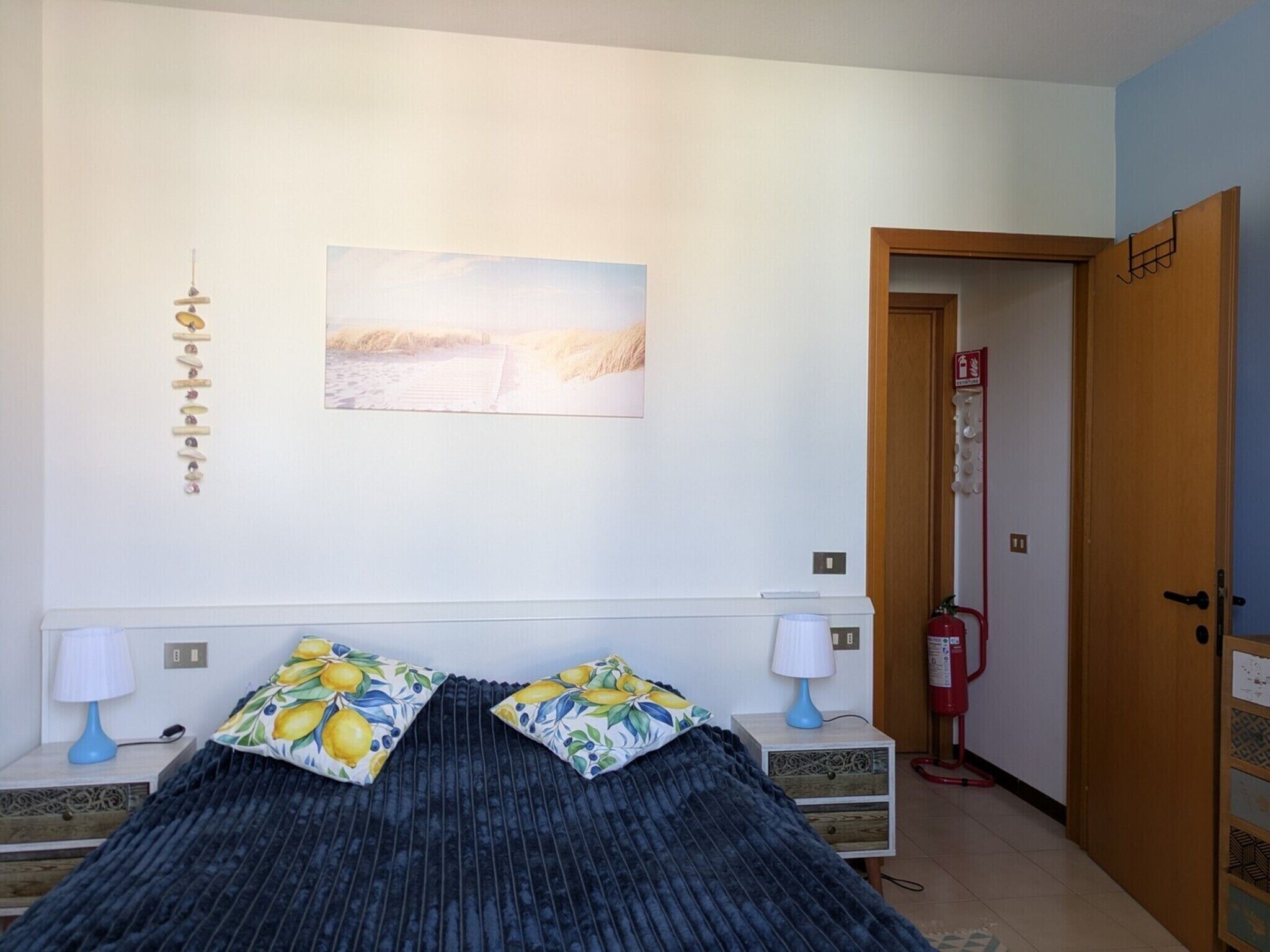 A bedroom with a bed, two bedside tables, a wall-mounted lamp, a painting, and a door leading to another room.