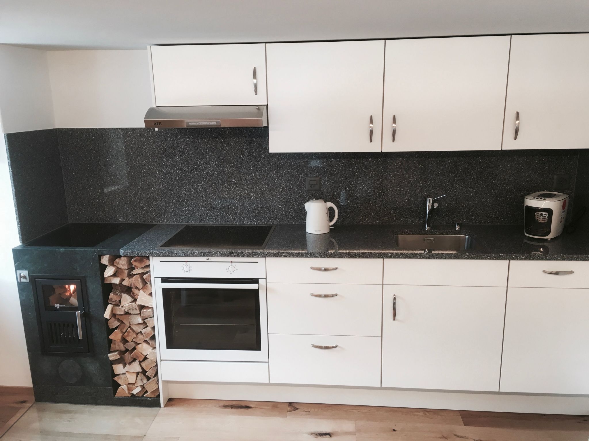A modern kitchen with white cabinets, a black countertop, a built-in oven, and a wood stove.