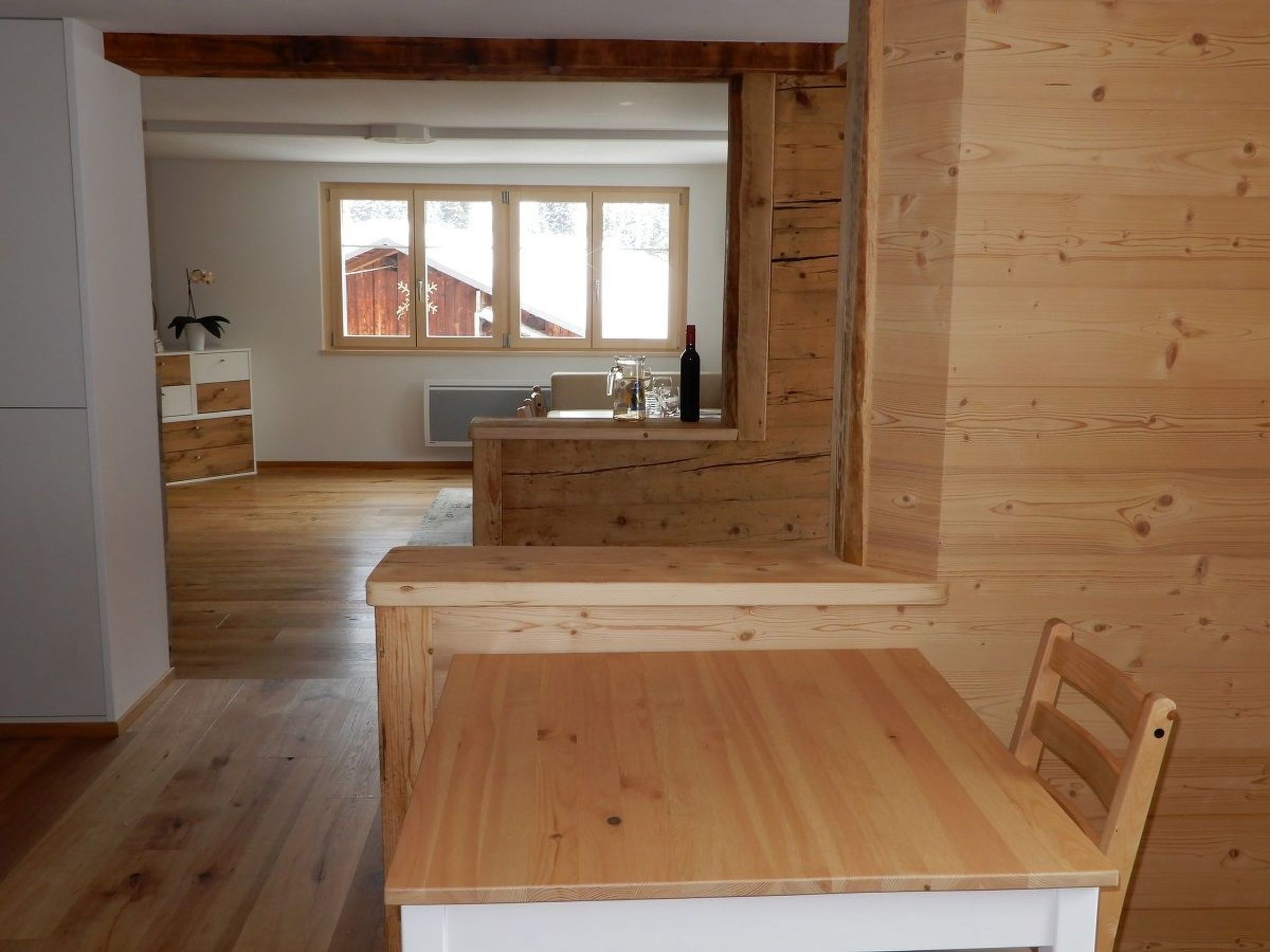 A wooden interior with a dining table and chairs, a kitchen area with a window, and a view of the outside.