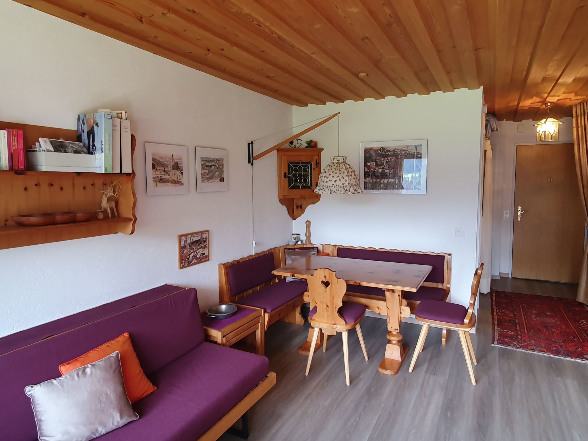 Une pièce chaleureuse avec un plafond en bois, un canapé violet, une table à manger en bois avec des bancs, et une petite étagère remplie de livres et de photos.