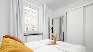 Une chambre à coucher avec un lit, une fenêtre, un radiateur, une porte et une petite table sur laquelle se trouvent une tasse et des fleurs.