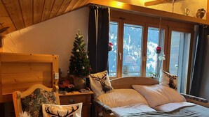 Ein gemütliches Zimmer mit einem Bett, einem Sessel mit einem Deer-Kissen, einer kleinen Weihnachtstanne und einem Fenster mit Blick auf schneebedeckte Berge.