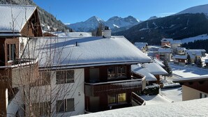 Ein verschneites Bergdorf mit traditionellen Holzhäusern, einem strahlend blauen Himmel und Blick auf schneebedeckte Gipfel im Hintergrund.