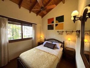 Une chambre à coucher avec un lit, un ventilateur de plafond, une fenêtre avec des rideaux, des œuvres d’art murales et des lampes de chevet.