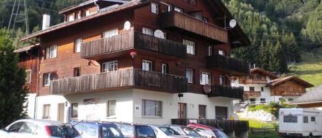A multi-story building with balconies, a parking lot with several cars, and a mountainous backdrop.