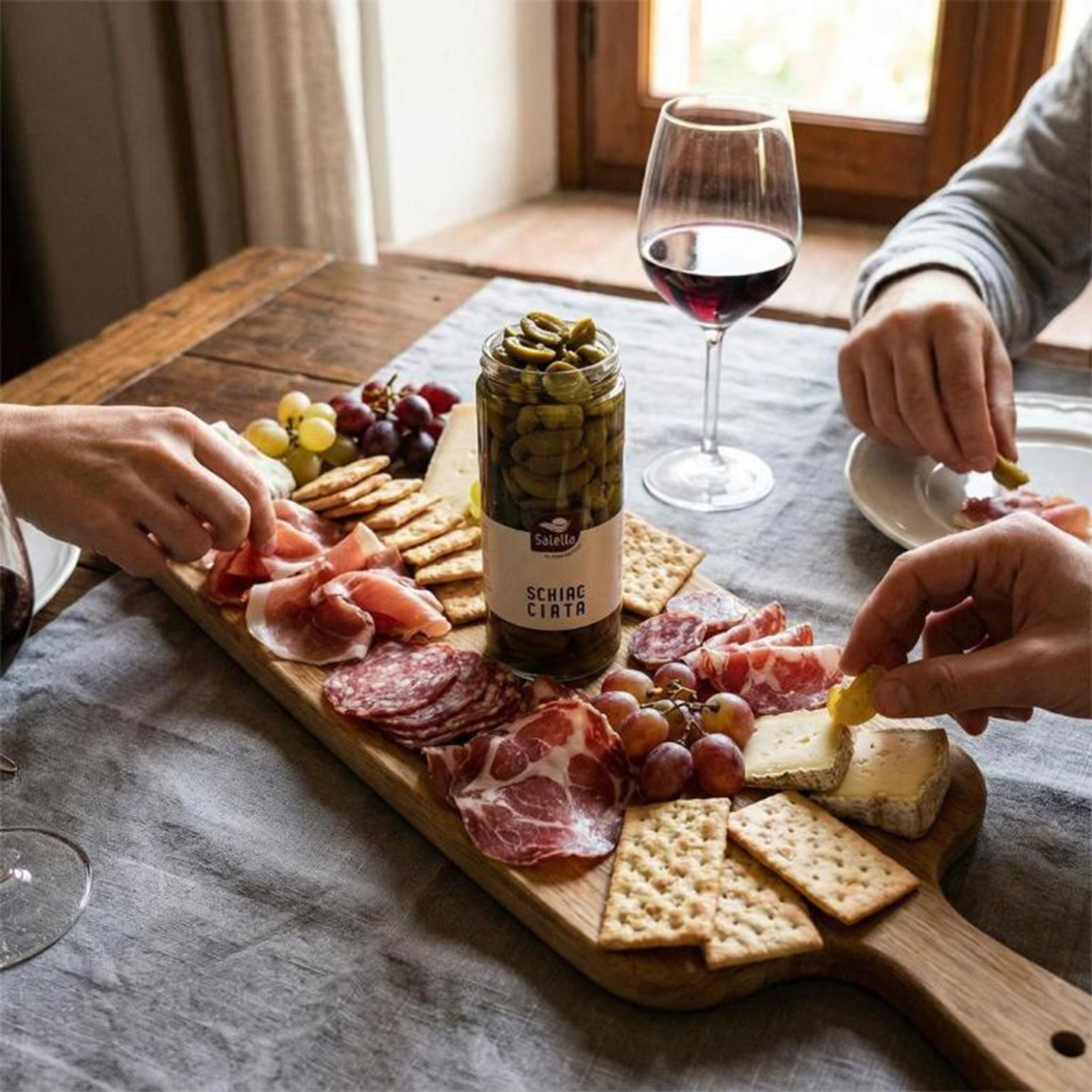 A wooden board with various cheeses, cured meats, and crackers, a bottle of Salsiccia C.IATA, and a glass of red wine on a table.