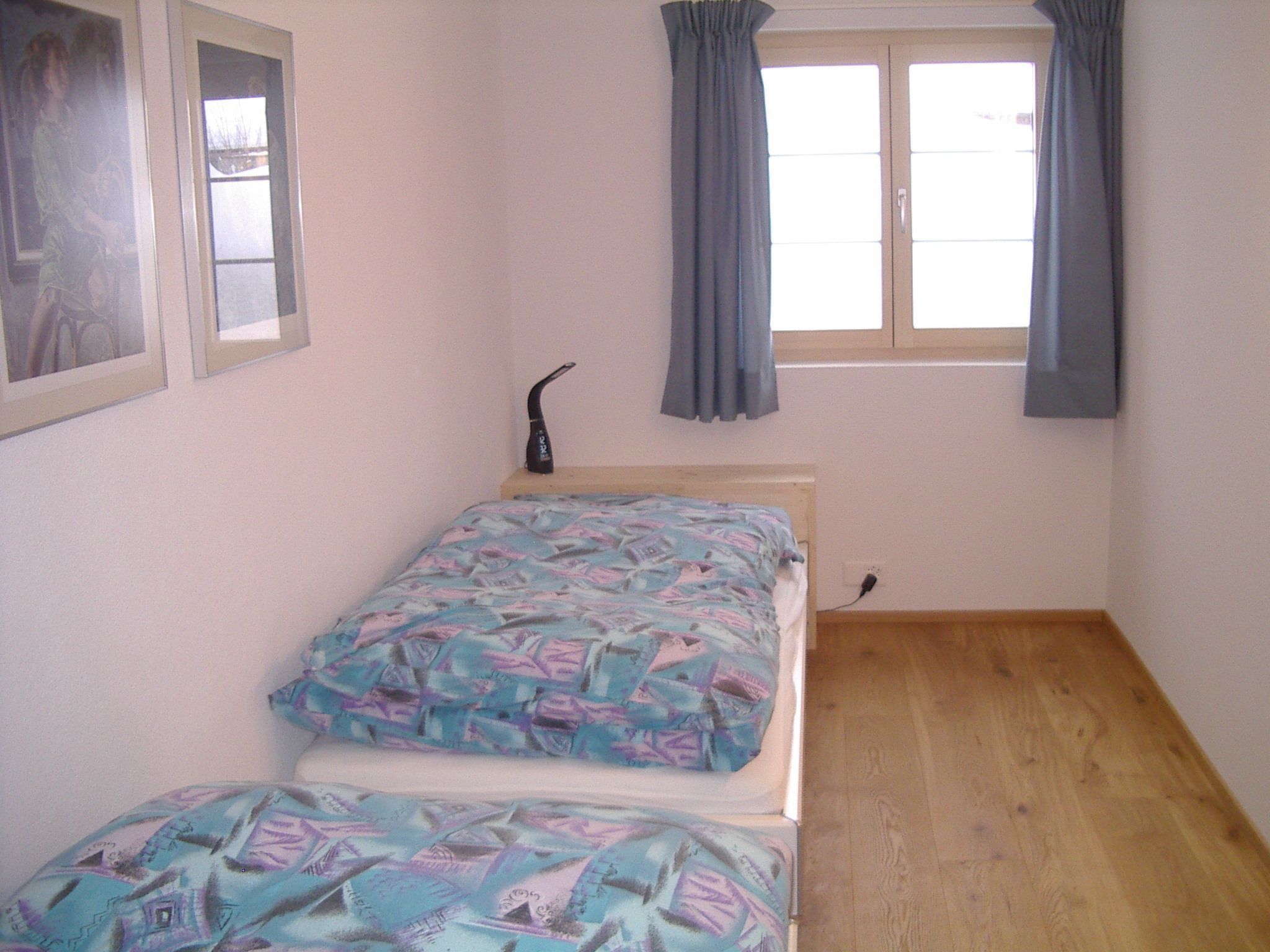 Een kleine kamer met een enkel bed, een nachtkastje, een raam met gordijnen en een wandradiator.