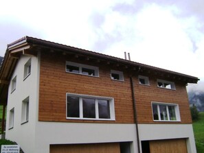 A two-story building with a wooden facade and white lower section, featuring multiple windows and a garage entrance. - Casa Aurora Hendry-Canepa, (Sedrun / Cavorgia), 610.01 by Interhome (Sedrun)