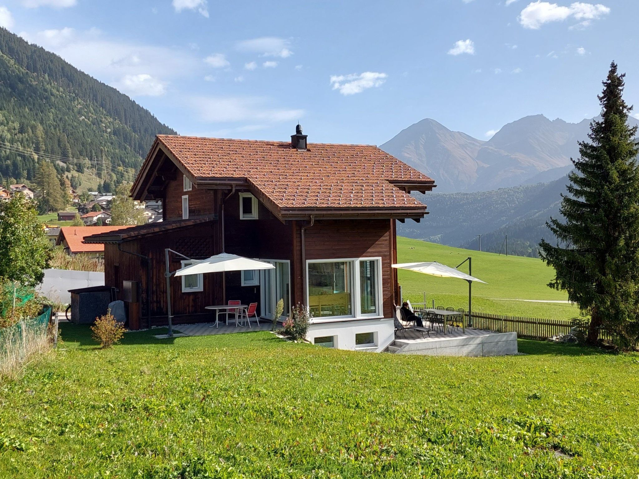 A wooden house with a red-tiled roof, a patio with outdoor furniture, and a view of mountains in the background.