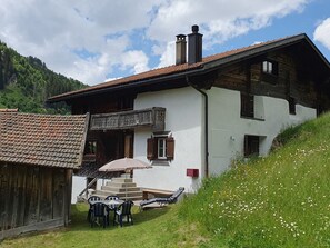 Un chalet à deux étages, doté d’un toit en pente, d’un balcon et d’un petit espace extérieur aménagé avec une table et des chaises.