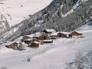 A snow-covered mountain village with wooden buildings, including a lodge with red accents, surrounded by snow-covered trees and hills.