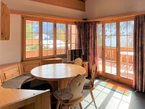 Une pièce comprenant une table à manger en bois, des chaises, un téléviseur et de grandes fenêtres avec des rideaux.