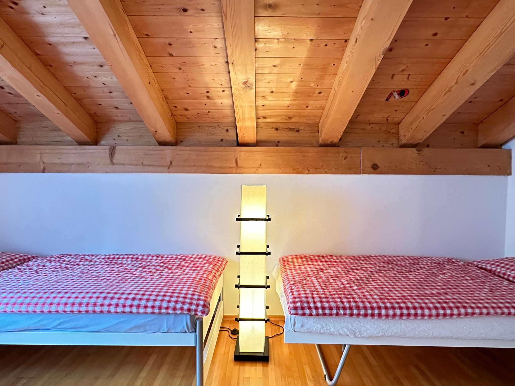 Une chambre avec deux lits, des poutres apparentes en bois au plafond et un luminaire fixé au mur.