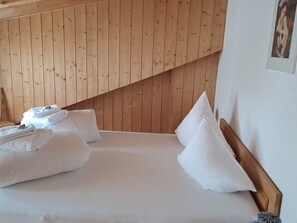 Cama bem arrumada com roupa de cama e almofadas brancas, cabeceira de madeira e teto inclinado de madeira.