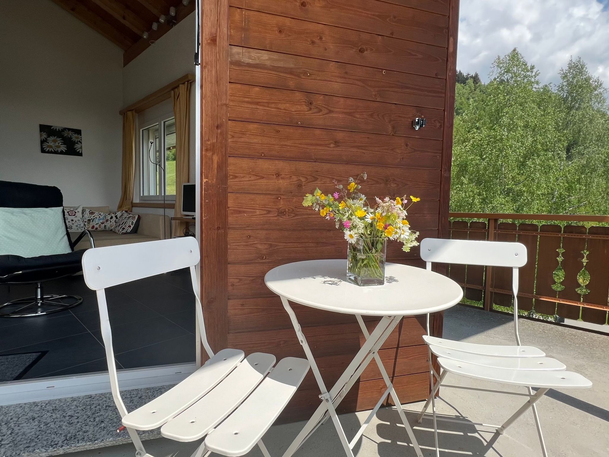 A wooden deck with a white table and chairs, a vase of flowers, and a view of greenery.