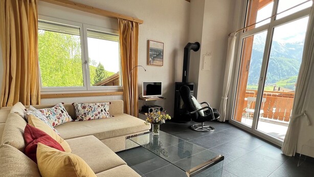 A living room with a sofa, a glass coffee table, a black chair, a fireplace, and a view of mountains through large windows. - Casa Diala Panorama, (Segnas/Disentis), 1106 by Interhome (Disentis)