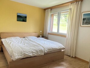 Ein Bett mit Holz-Kopfstütze, ein Nachttisch mit Lampe, ein Fenster mit Vorhängen und ein Bild an der Wand.