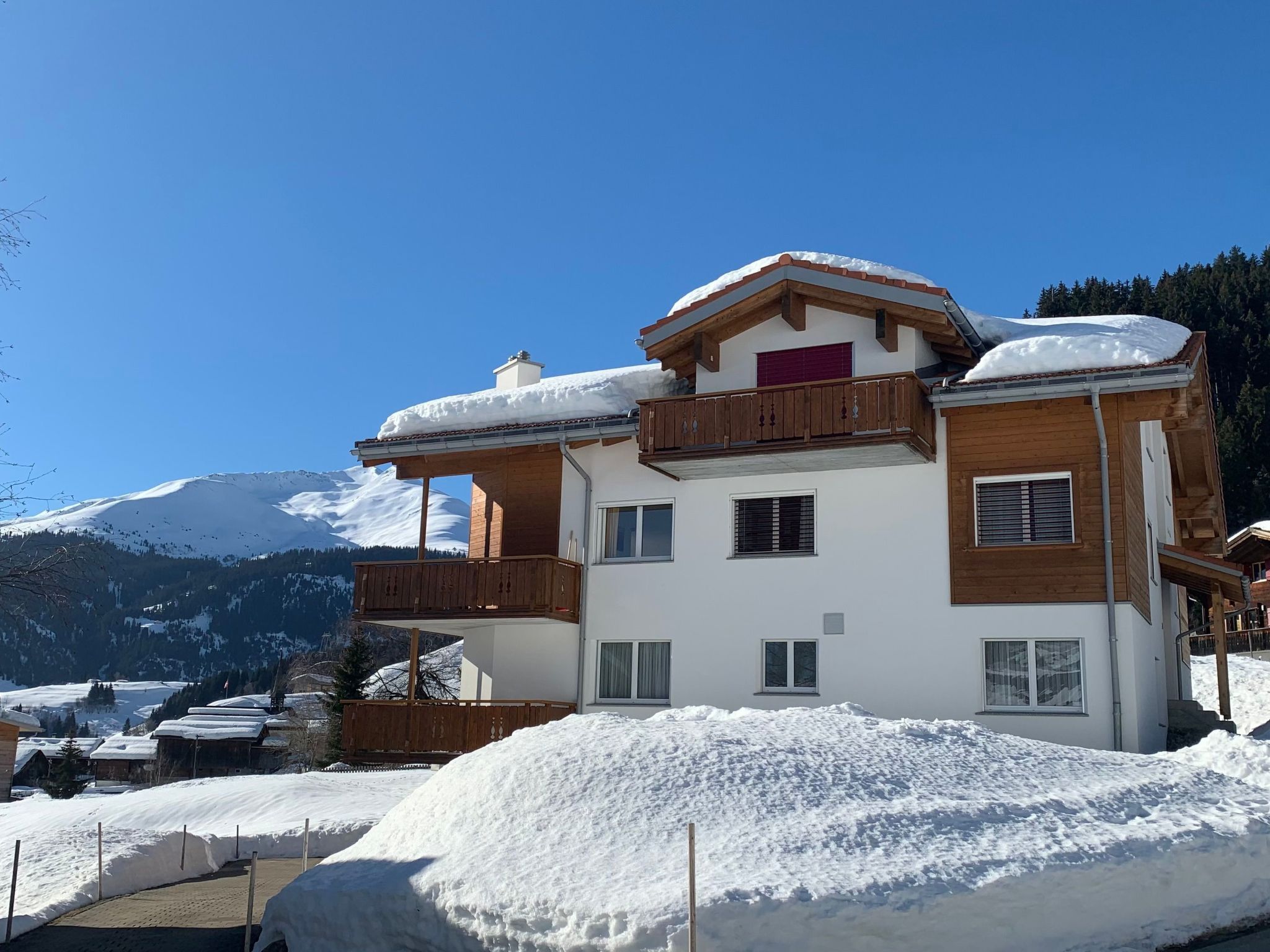 A two-story chalet with a balcony, surrounded by snow-covered mountains.