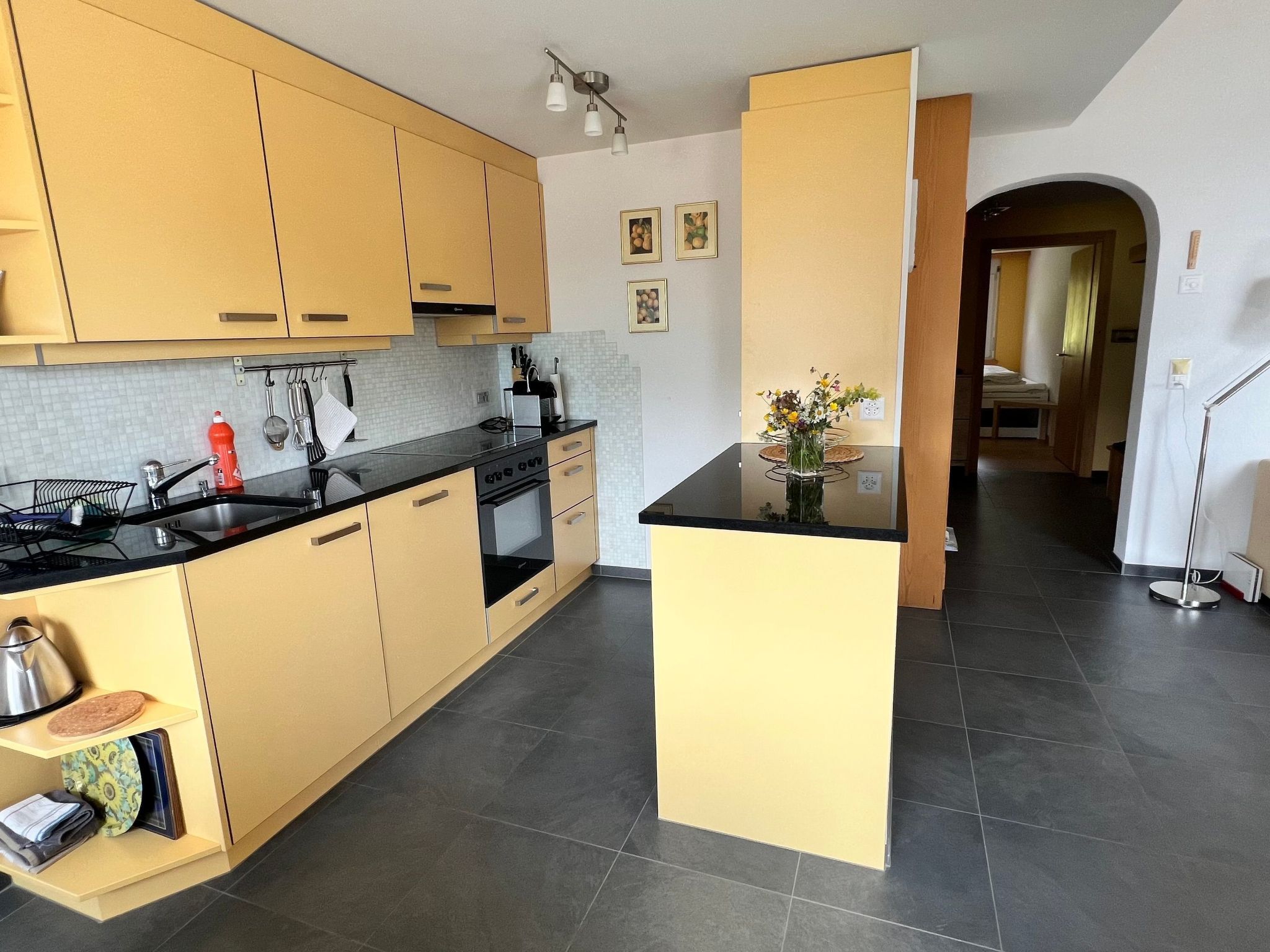 A modern kitchen with light-colored cabinets, a black countertop, and a central island. There is a sink, stove, and various kitchen utensils visible.
