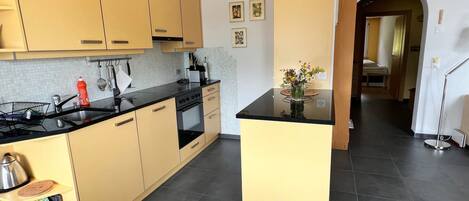 A modern kitchen with light-colored cabinets, a black countertop, and a central island. There is a sink, stove, and various kitchen utensils visible.