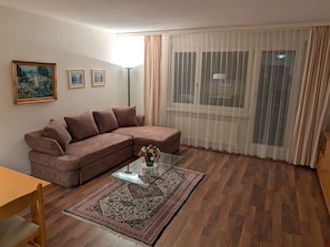 A living room with a brown sofa, a glass coffee table with a vase, a patterned rug, and a large window with sheer curtains. - Disentiserhof Haus C 102, (Disentis/Mustér), 1038 by Interhome (Disentis)