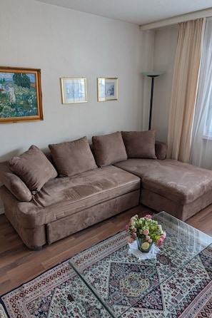 A living room with a brown sectional sofa, two framed pictures on the wall, a floor lamp, and a glass coffee table with a floral arrangement. - Disentiserhof Haus C 102, (Disentis/Mustér), 1038 by Interhome (Disentis)