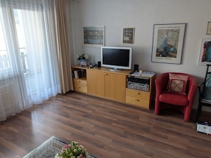 Een woonkamer met een flatscreen televisie, houten kasten, een rode fauteuil en een raam met dunne gordijnen.