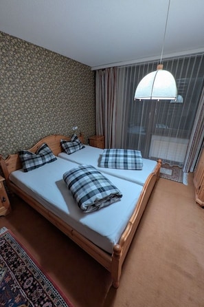 A double bed with a wooden frame, two pillows, a small bedside table, a patterned rug, and a window with curtains. - Disentiserhof Haus C 102, (Disentis/Mustér), 1038 by Interhome (Disentis)