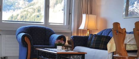 En stue med udsigt til bjerge, en sofa, en lænestol, et bord med en vase og en lampe.