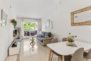 Ein modernes Wohnzimmer mit einer Couch, einem Esstisch, Stühlen, einem Balkon mit Tisch und Stühlen sowie einem großen, gerahmten Bild an der Wand.