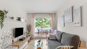 Ein modernes Wohnzimmer mit Flachbildfernseher, einem Glastisch, einer Couch und einem Balkon mit Gartenmöbeln.