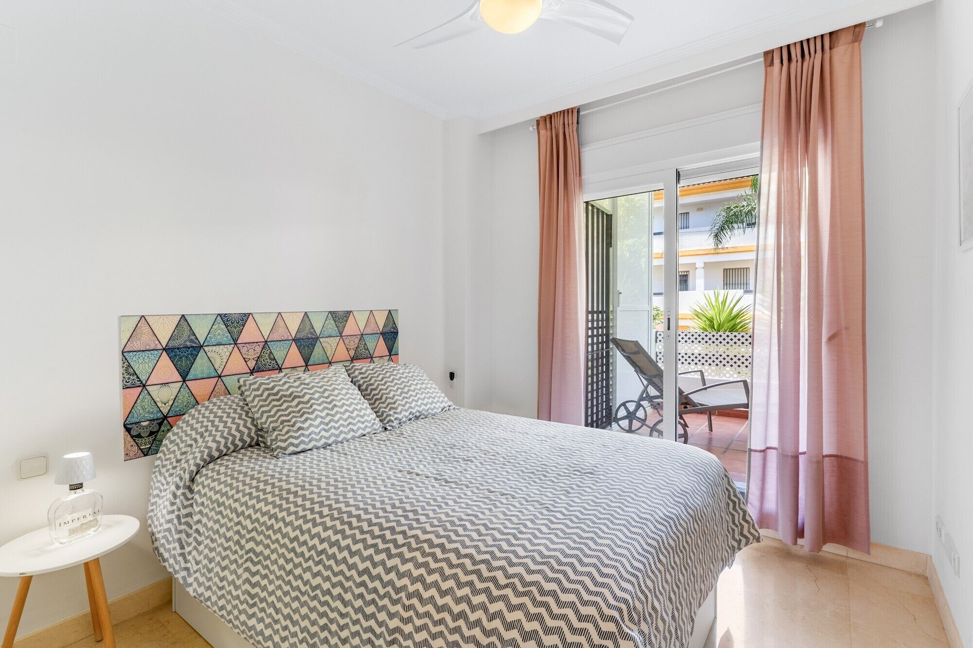 A bedroom with a bed, a headboard with a geometric pattern, a small white table with a bottle, and a view of an outdoor area with a table and chairs.
