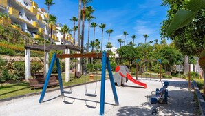 Ein Spielplatz mit Schaukeln und einer Rutsche, umgeben von Palmen und einem Gebäude mit Balkonen.
