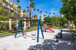 Ein Spielplatz mit Schaukeln und einer Rutsche, umgeben von Palmen und einem Gebäude mit Balkonen.