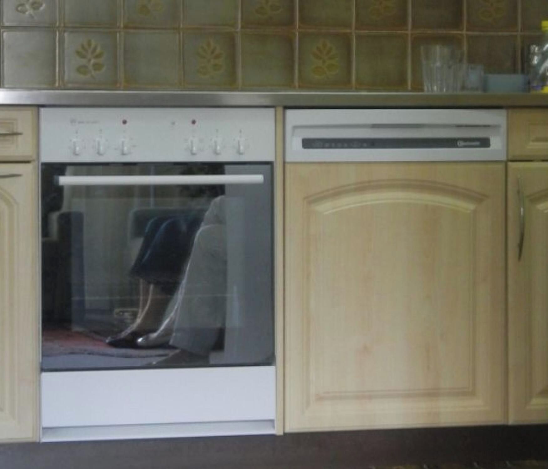 Une cuisine équipée d’un four encastré et d’un lave-vaisselle, avec des meubles en bois et un dosseret carrelé.