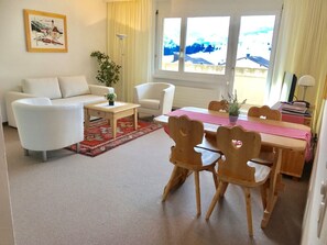 Ein Wohnzimmer mit Sofa, Couchtisch, Esstisch mit Stühlen, einem Fenster mit Vorhängen und einem Bild an der Wand.